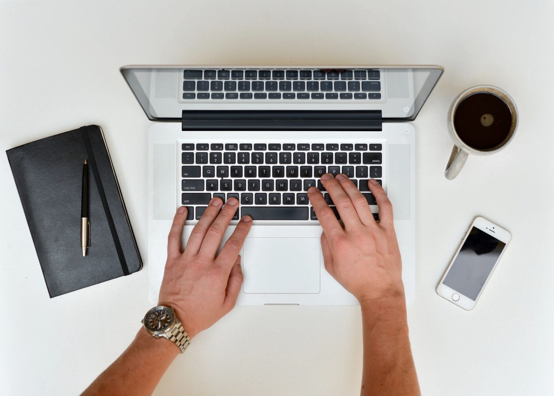 Overhead view of hands typing on a laptop with a notebook, coffee, and smartphone on a white desk.