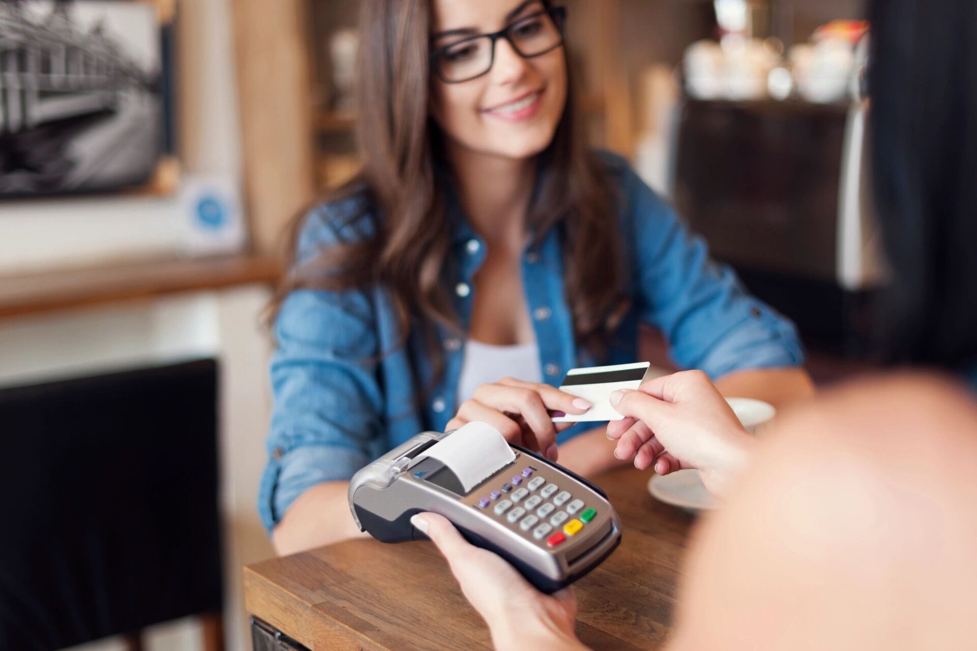 Smiling woman handing a credit card to a barista holding a payment terminal inside a café.