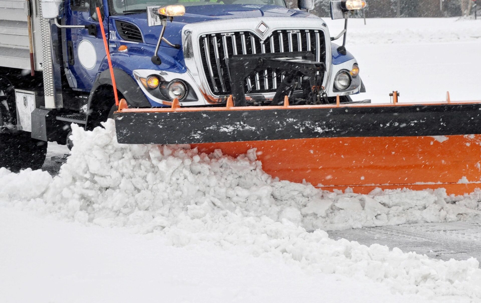 Snowplow truck with orange blade pushing snow off a city road during winter weather.