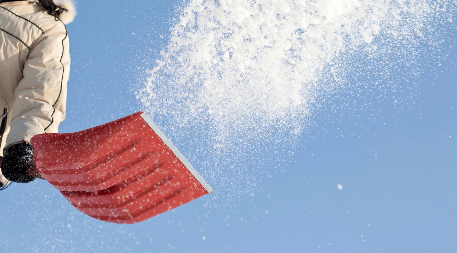 Red snow shovel tossing a spray of snow into the air against a clear blue sky.