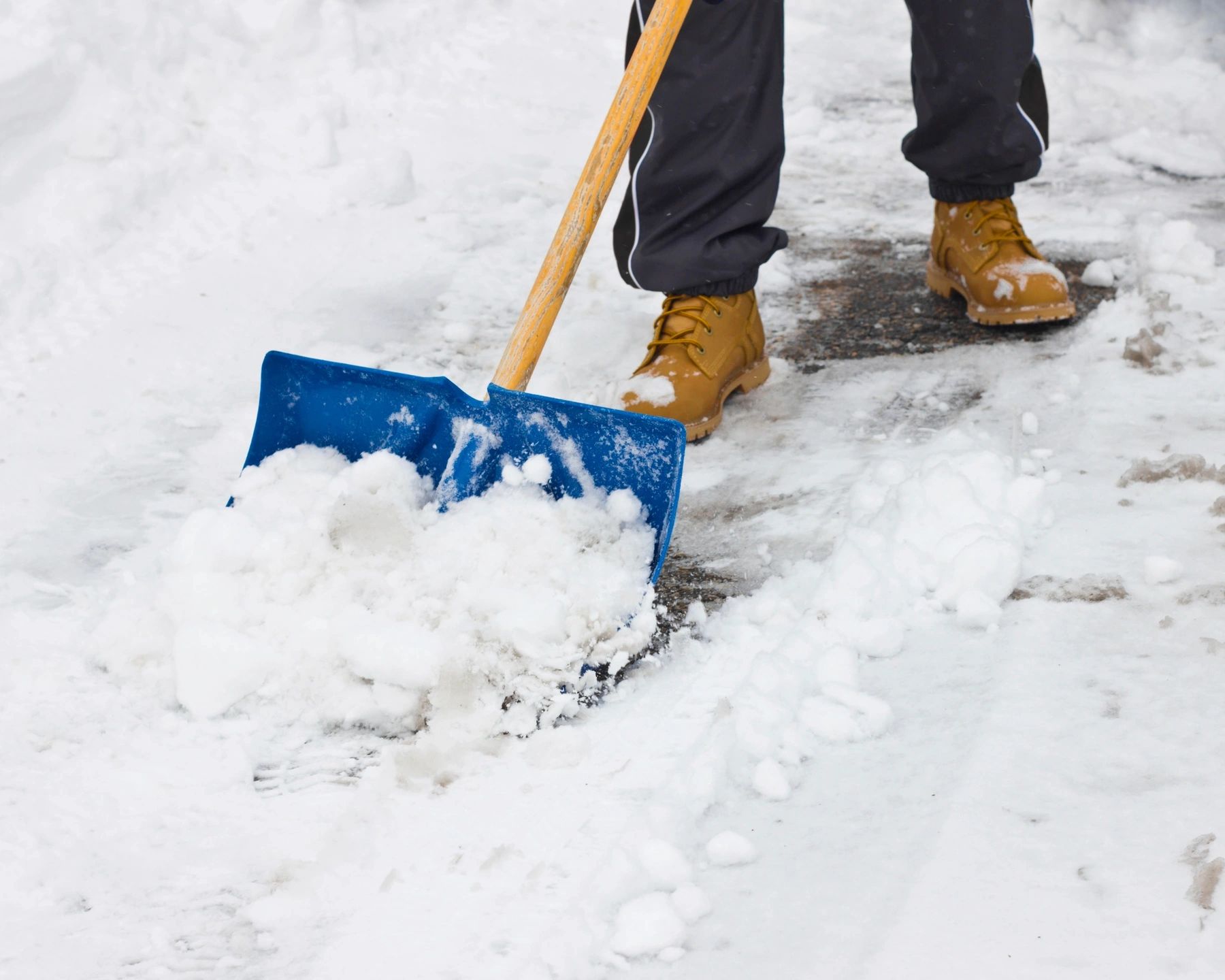 Person shoveling snow with a blue snow shovel, wearing boots and winter pants on a snowy sidewalk.